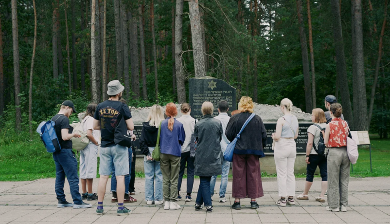Gruppe junger Menschen und Fachkräfte bei einem Besuch einer Gedenkstätte. Sie stehen vor einem Denkmal in einer Waldlandschaft und beschäftigen sich gemeinsam mit Erinnerungskultur und historischer Bildung.