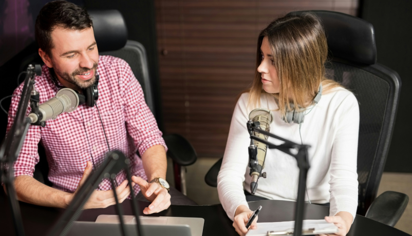 Zwei Personen mit Headsets sprechen in einem Radiostudio an einem Tisch vor Mikrofonen