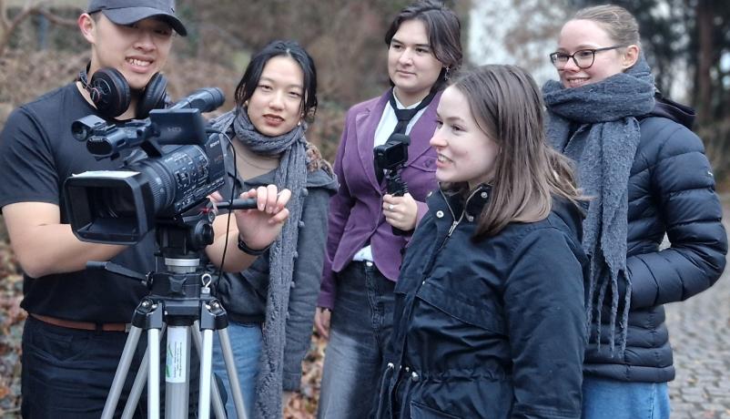 Gruppe junger Menschen bei einer Filmaufnahme im Freien; ein Teilnehmer bedient eine Videokamera