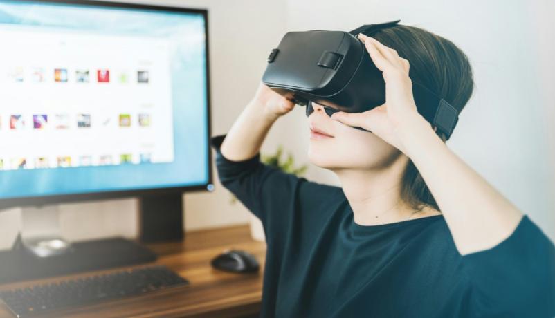Eine Frau trägt ein schwarzes VR-Headset und steht neben einem Cmputerbildschirm