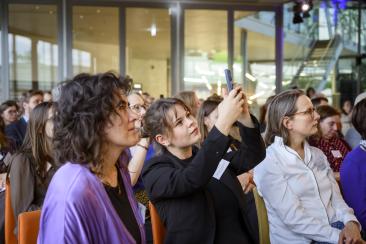 Gäste verfolgen das Programm; eine Person fotografiert mit dem Smartphone.