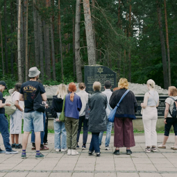 Gruppe junger Menschen und Fachkräfte bei einem Besuch einer Gedenkstätte. Sie stehen vor einem Denkmal in einer Waldlandschaft und beschäftigen sich gemeinsam mit Erinnerungskultur und historischer Bildung.